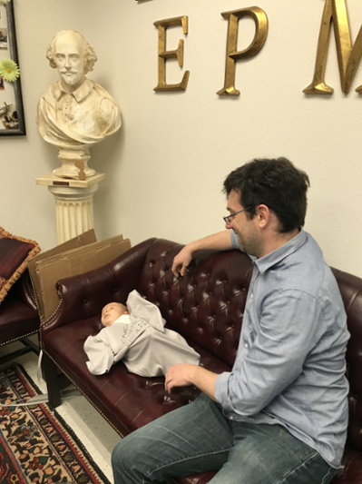 A small baby, wearing an EPML t-shirt, lies on the couch in the EPML lounge, alongside faculty member Matthew Harrison